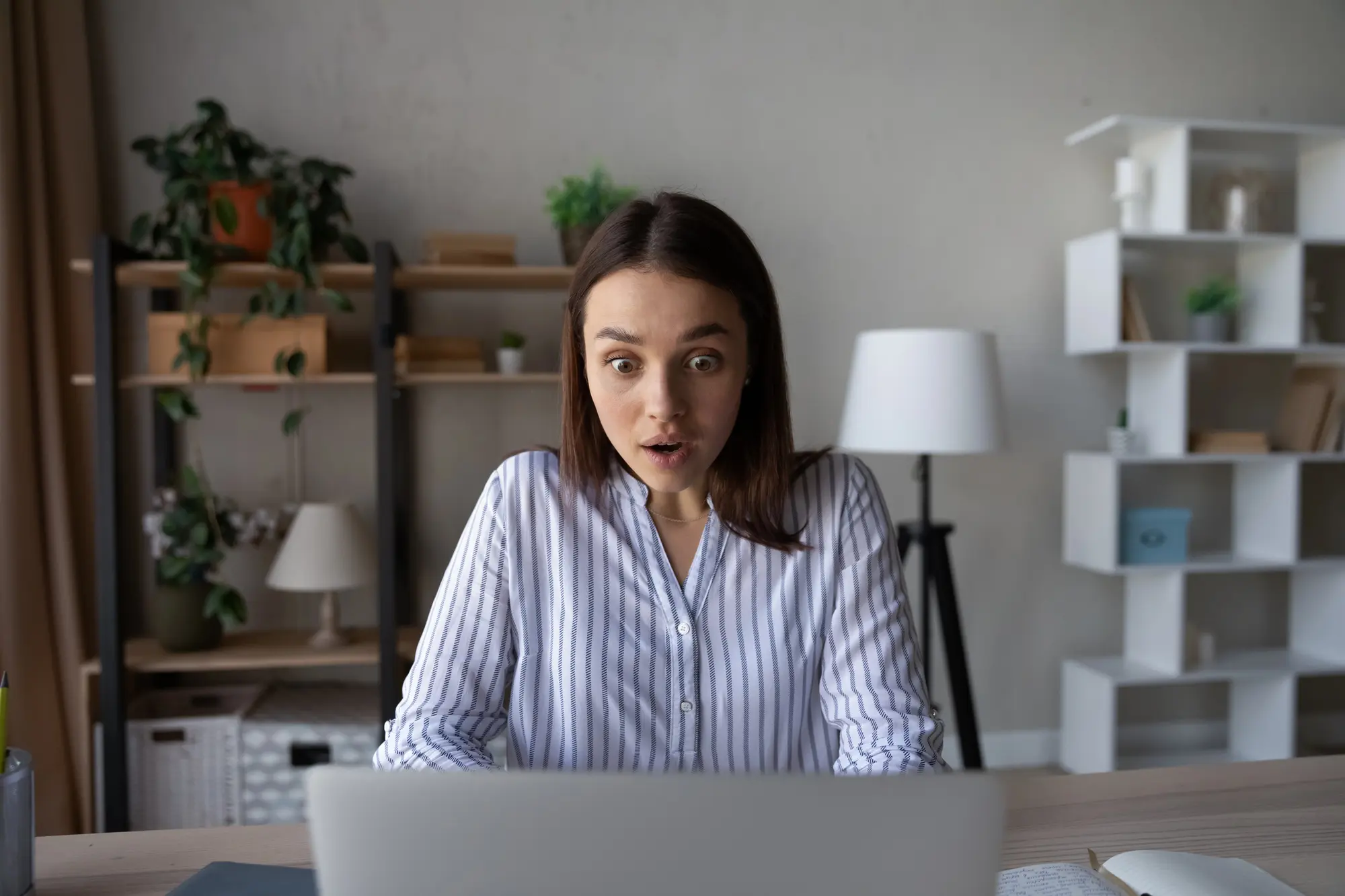 Een vrouw met steil bruin haar en een gestreept shirt zit aan een bureau en kijkt met een verbaasde blik naar het scherm van een laptop omdat haar WordPress website is gehackt.